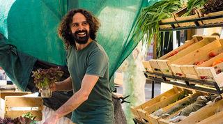 Smiling, bearded man arranges vegetables at an open market stall